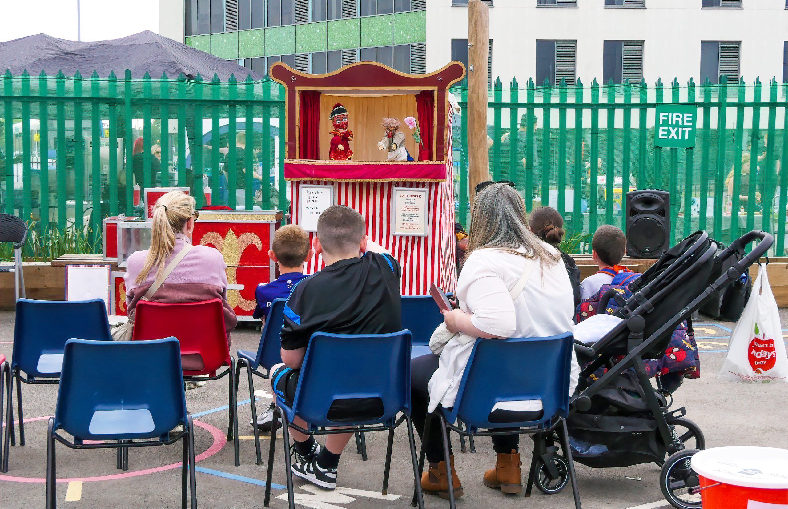 Audience seated in blue and red chairs watching a red-and-white striped puppet booth with two puppets performing on a small stage behind a green fence.