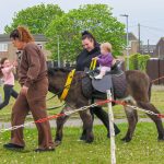Child riding a pony with a supervising adult at an outdoor event, grassy field and spectators in the background.