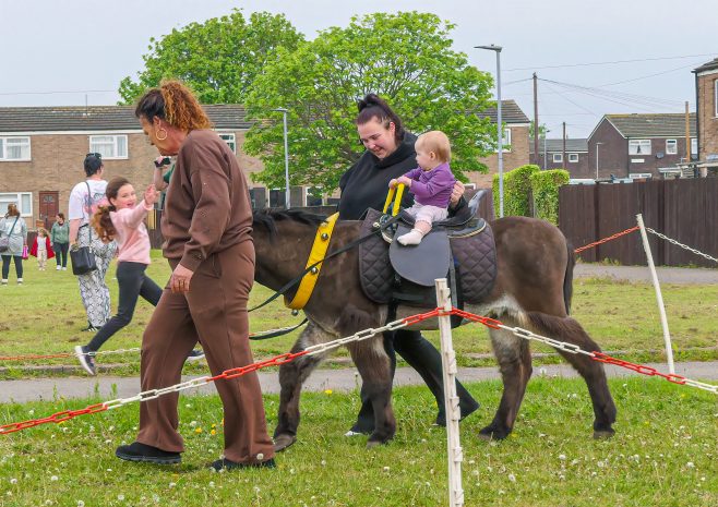 Child riding a pony with a supervising adult at an outdoor event, grassy field and spectators in the background.