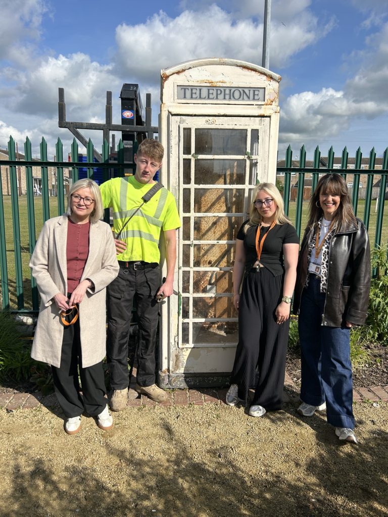 Four adults stand together beside a weathered white telephone booth behind a green metal fence on a sunny day in a park-like setting.