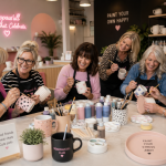 Six women around a table painting ceramic mugs at a colorful workshop, smiles and craft supplies visible beneath pink decor in the background.