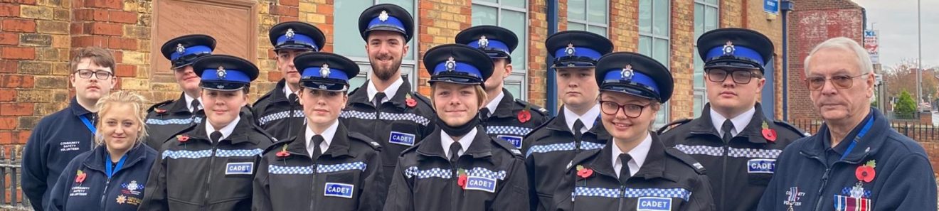 Group of cadets in blue-and-black ceremonial uniforms and peaked caps pose outside a brick building, flanked by two adult volunteers wearing badges and poppy pins.