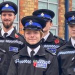 Group of cadets in blue-and-black ceremonial uniforms and peaked caps pose outside a brick building, flanked by two adult volunteers wearing badges and poppy pins.