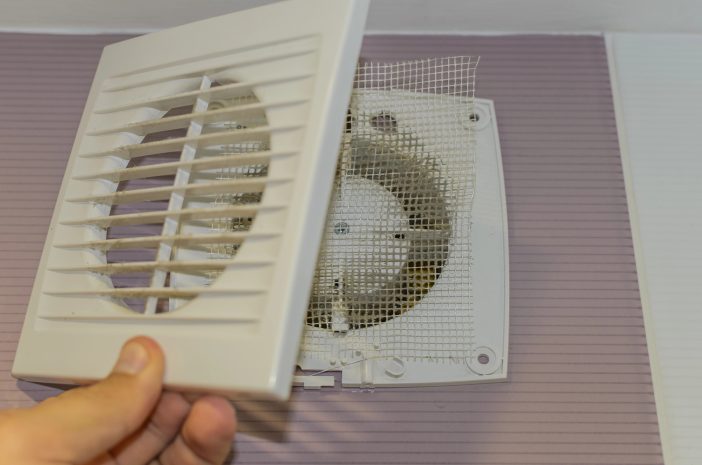 Person removing a white louvered vent cover from an indoor air vent, exposing dust and interior mesh