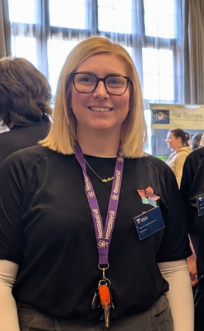 Smiling young woman with blonde hair and glasses, wearing a black shirt and purple lanyard with a badge at an indoor event.