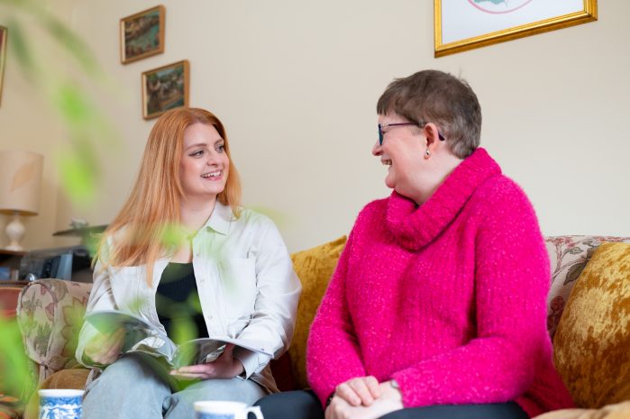 Two women sit on a floral sofa, smiling and chatting in a cozy living room, one holding magazines/papers in her lap.
