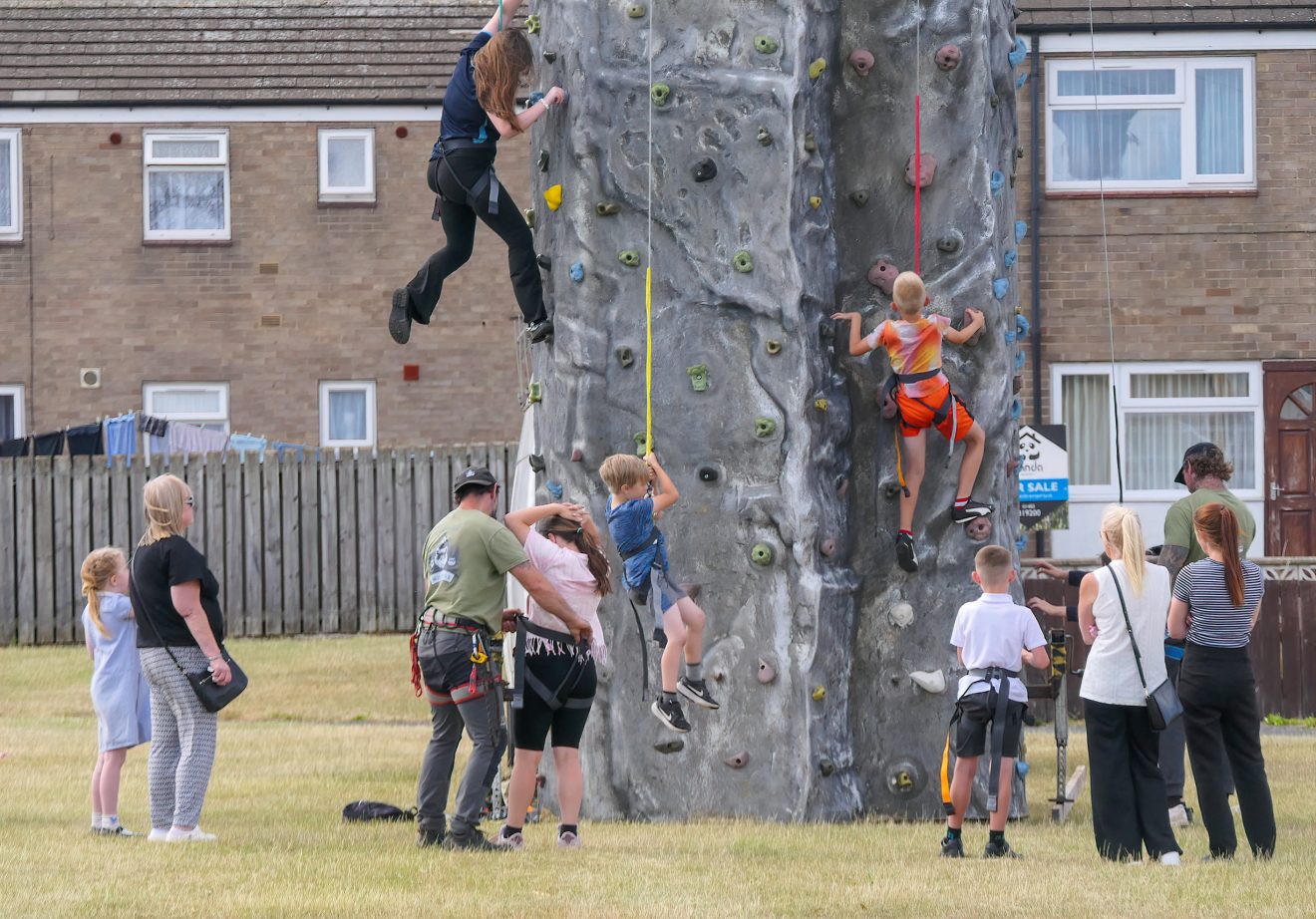 Children and adults in harnesses watch a climber on a tall outdoor artificial rock wall in a yard or park.