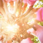 Group of people in pink shirts join hands in a circle, showing teamwork outdoors.